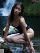 A woman in a black bathing suit sitting on a rock by a waterfall.